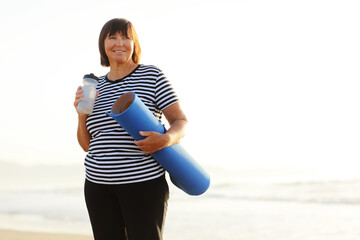 Smiling mature adult woman with bottle of water after exercising outdoors holding yoga mat outdoors on beach by sea on summer. Senior healthy lifestyle, vitality, healthcare concept. copy space