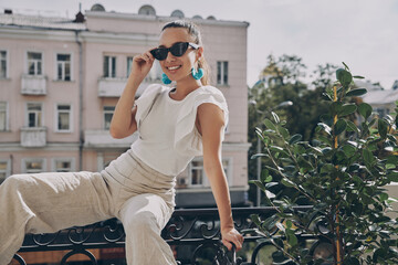 Naklejka na ściany i meble Stylish young woman adjusting her eyeglasses and smiling while sitting on the balcony