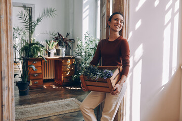 Happy young woman carrying wooden crate with plants while leaning at the doorway at home