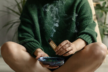 faceless middle-aged woman in a green pullover holds a wooden palo santo stick with a magical stream of smoke before meditation and yoga sitting on the floor of the house