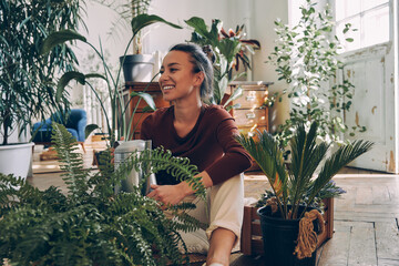 Happy young woman watering houseplants while sitting on the floor at home