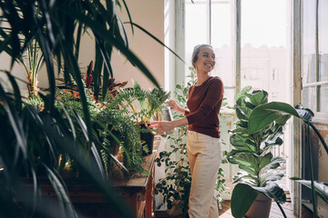 Confident young woman watering houseplants and smiling while standing at the domestic room