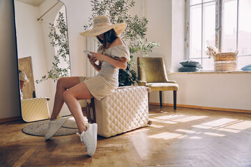 Attractive young woman in elegant hat sitting on suitcase and using smart phone