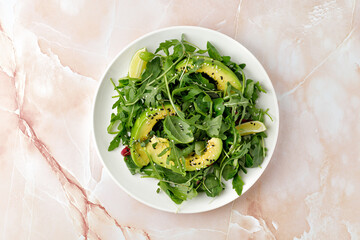 Fresh vegetable green salad with avocado and mix lettuce, arugula on white plate and beige marble background.