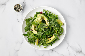 Green healthy salad with avocado and mix lettuce, arugula on white plate and marble background. Top...
