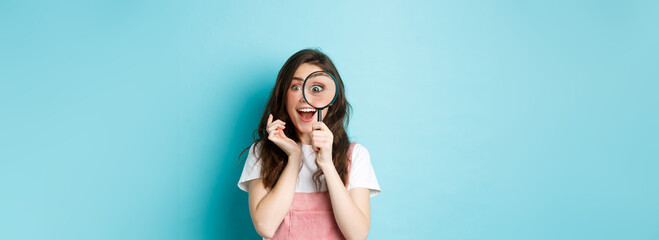 Happy young woman looking through magnifying glass with excited face, found or search something, standing over blue background - obrazy, fototapety, plakaty