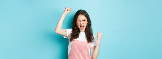 Cheerful young woman rooting for team, watching sports game and cheering, raising hand up in fist...
