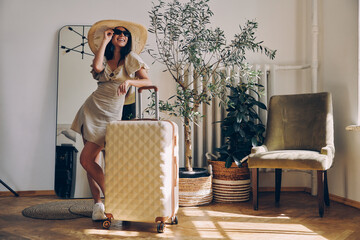 Dreamful young woman in elegant hat leaning on the suitcase and smiling while standing at home