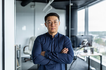 Portrait of serious Asian developer, businessman with crossed arms looking concentrated at camera, man in glasses and shirt working inside modern office building.
