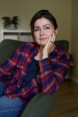 Fototapeta na wymiar Vertical portrait positive lonely pretty and thoughtful woman in plaid shirt looking at camera and sitting in armchair.