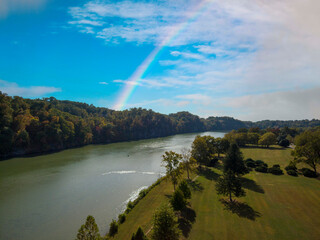 an aerial shot of a gorgeous autumn landscape along the silky green waters of the Tennessee River surrounded by autumn colored trees and lush green trees with blue sky, clouds and a rainbow - obrazy, fototapety, plakaty