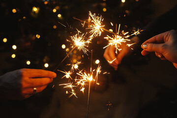 Happy New Year! Friends celebrating with burning sparklers in hands against christmas tree lights...