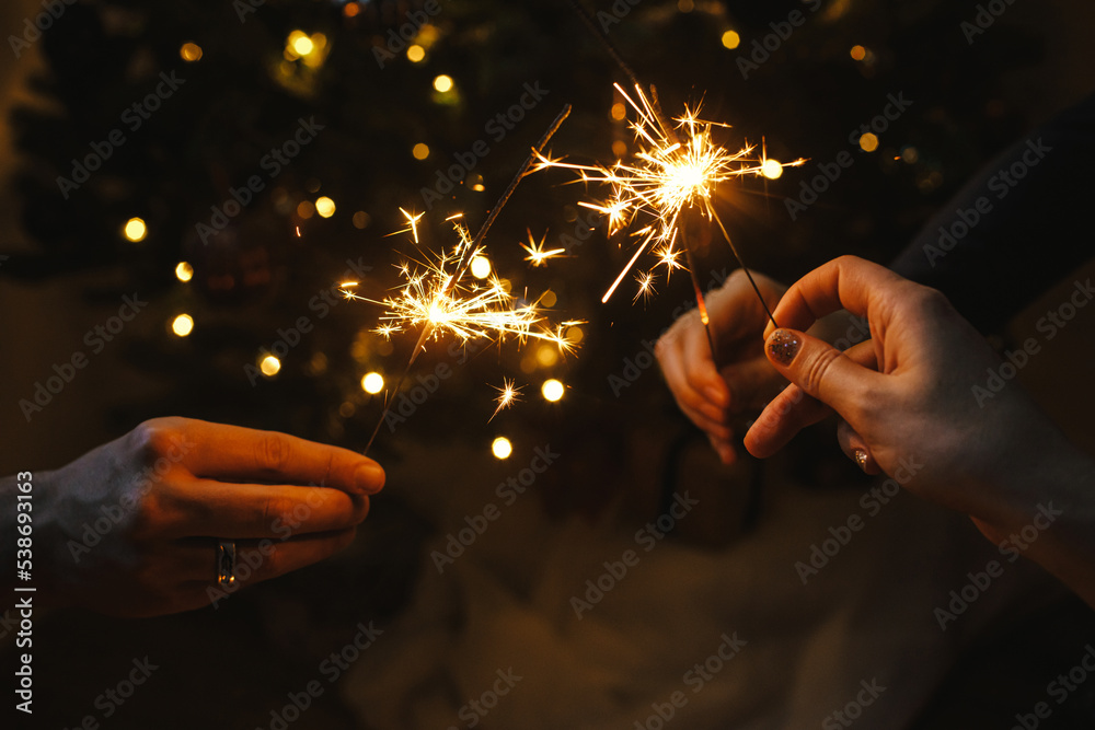 Wall mural Happy New Year! Friends celebrating with burning sparklers in hands against christmas tree lights in dark room. Hands holding fireworks on background of stylish decorated illuminated tree. Moody