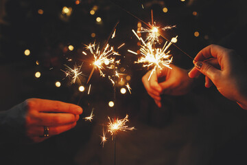 Happy New Year! Friends celebrating with burning sparklers in hands against christmas tree lights in dark room. Hands holding fireworks on background of stylish decorated illuminated tree. Moody - Powered by Adobe