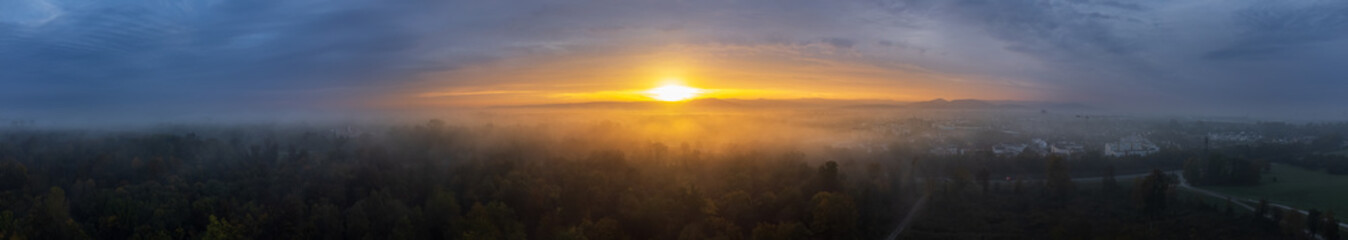 Panoramic view over a forest and a city on a foggy autumn morning while the sun rises in the background