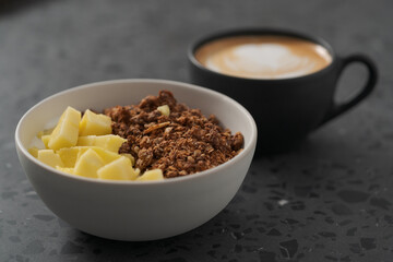 chocolate granola with mango and greek yogurt in white bowl with black cup with cappuccino on concrete background