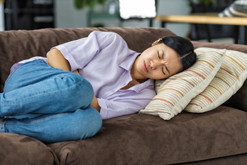 Young Asian woman suffering from abdominal pain lying on the couch at home. Female suffering from...