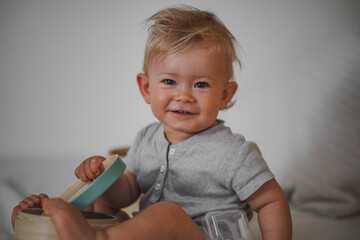 Naklejka premium cute happy one year old blond baby boy in casual outfit at home on a cozy sofa smiling or looking into the Camera while open can or box