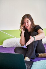 Vertical shot of a young woman sitting on a bed with a laptop and happily talking on a phone