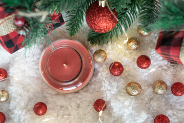 Red candle in glass and gifts in a checkered cloth wrapper, yellow lights on a garland, a sprig of a Christmas tree on a white soft background. Top view