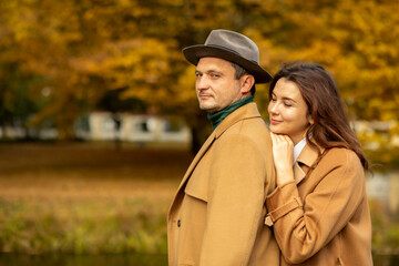 Happy couple (wife and husband): woman hugging man from back. Orange, yellow autumn park is on background,love concept,happy lifestyle. Stylish adults wear warm beige coats and hat. Copy space on left