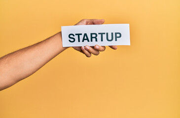 Hand of caucasian man holding paper with startup word over isolated yellow background