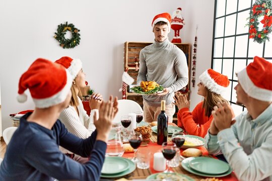 Group of people meeting clapping and sitting on the table. Man standing and holding roasted turkey celebrating Christmas at home.