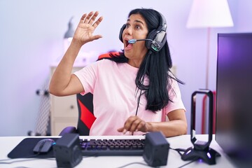 Mature hispanic woman playing video games at home shouting and screaming loud to side with hand on mouth. communication concept.
