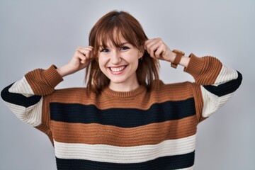 Young beautiful woman wearing striped sweater over isolated background smiling pulling ears with...
