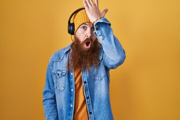 Caucasian man with long beard listening to music using headphones surprised with hand on head for...