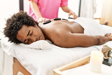 Obraz na płótnie Canvas Young african american man having massage using hot stones at beauty center