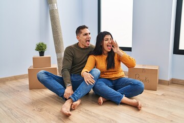 Fototapeta premium Young couple sitting on the floor at new home shouting and screaming loud to side with hand on mouth. communication concept.