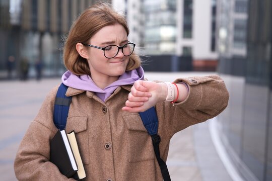 Stressed anxious young girl, woman is late busy university or college student with books, textbooks in glasses looking at her wrist watch, checking time in a hurry, rushing to lessons, exam. No time