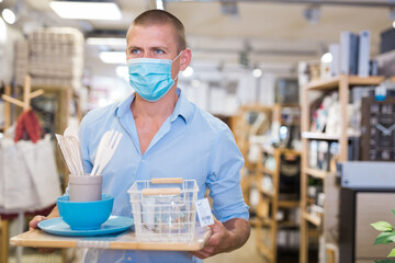 Young man client buyer in face mask choosing variety accessories and tableware in modern shop for...