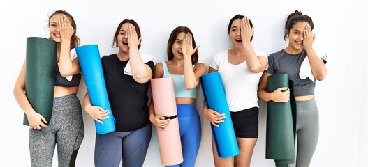 Group of women holding yoga mat standing over isolated background covering one eye with hand, confident smile on face and surprise emotion.