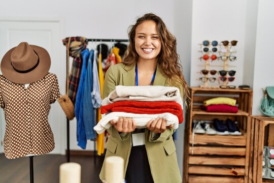 Young hispanic shopkeeper woman smiling happy holding stack of sweater at clothing store.