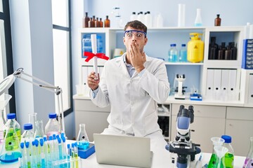 Young hispanic man working at scientist laboratory holding diploma covering mouth with hand, shocked and afraid for mistake. surprised expression