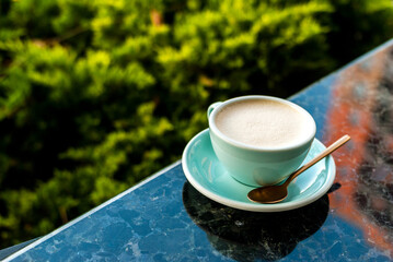 Cup of cappuccino on a marble table next to green spaces. Blue cup with coffee and a teaspoon on a...