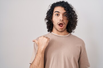 Hispanic man with curly hair standing over white background surprised pointing with hand finger to...