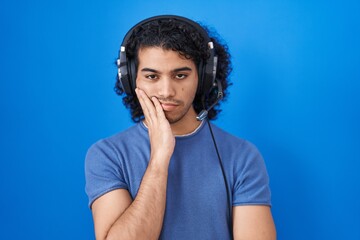Hispanic man with curly hair listening to music using headphones thinking looking tired and bored with depression problems with crossed arms.
