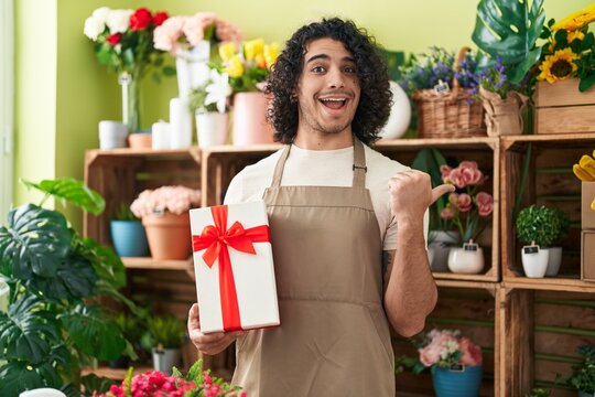 Hispanic man with curly hair working at florist shop holding gift pointing thumb up to the side smiling happy with open mouth