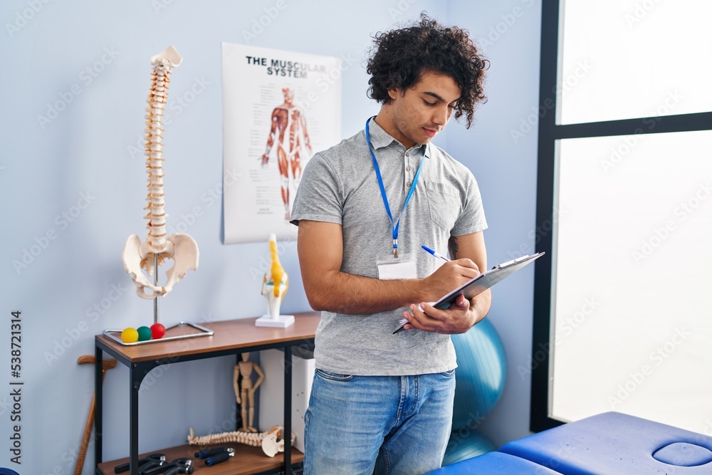 Sticker young hispanic man physiotherapist writing on clipboard at rehab clinic