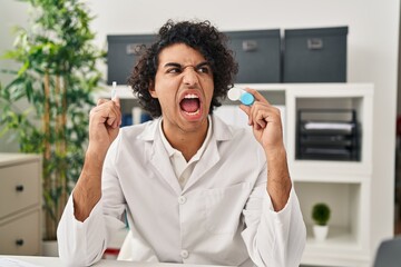 Hispanic man with curly hair holding contact lenses angry and mad screaming frustrated and furious, shouting with anger. rage and aggressive concept.