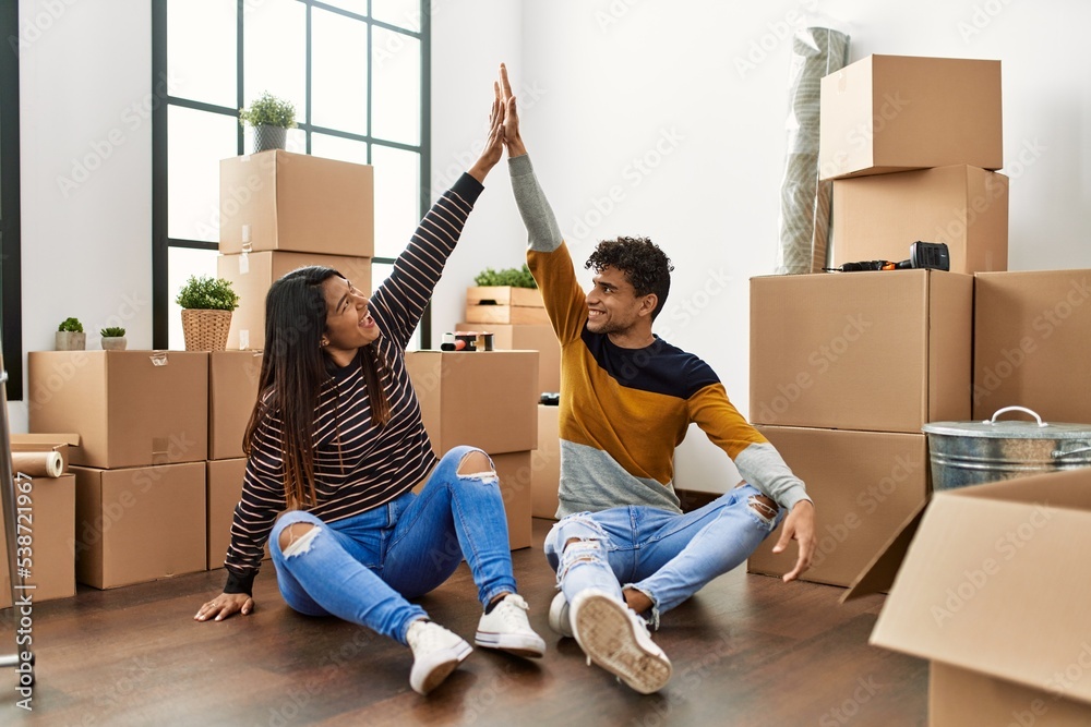 Poster young latin couple high five with hands raised up sitting on the floor at new home.