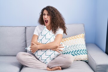 Hispanic woman with curly hair sitting on the sofa at home angry and mad screaming frustrated and...