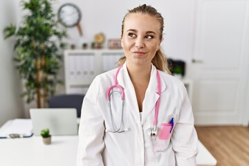 Young caucasian woman wearing doctor uniform and stethoscope at the clinic smiling looking to the side and staring away thinking.