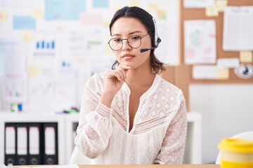 Hispanic young woman working at the office wearing headset and glasses thinking concentrated about...