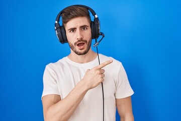 Hispanic man with beard listening to music wearing headphones surprised pointing with finger to the side, open mouth amazed expression.