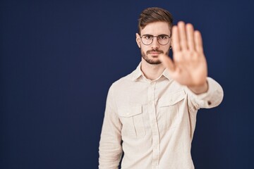 Hispanic man with beard standing over blue background doing stop sing with palm of the hand. warning expression with negative and serious gesture on the face.