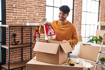 Young hispanic man smiling confident holding sneakers unpacking cardboard box at new home